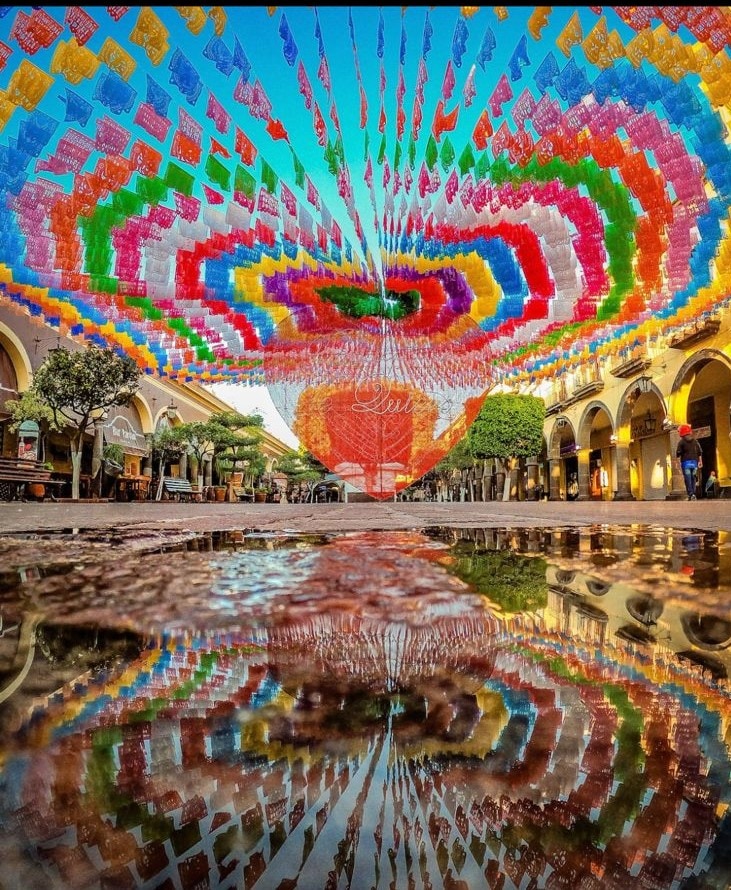 A vibrant street scene is depicted, showcasing colorful papel picado banners suspended above. The reflection of the decorations can be seen in a puddle on the cobblestone street, enhancing the festive atmosphere. Archways and trees line the street, contributing to the lively ambiance.