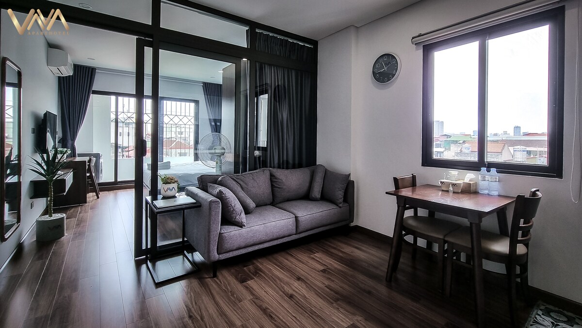 A modern living area features a spacious gray sofa positioned beside a wooden dining table with four chairs. Large windows allow natural light to fill the space, while a round clock adorns the wall. A view of the city is visible through the glass doors leading to the bedroom.