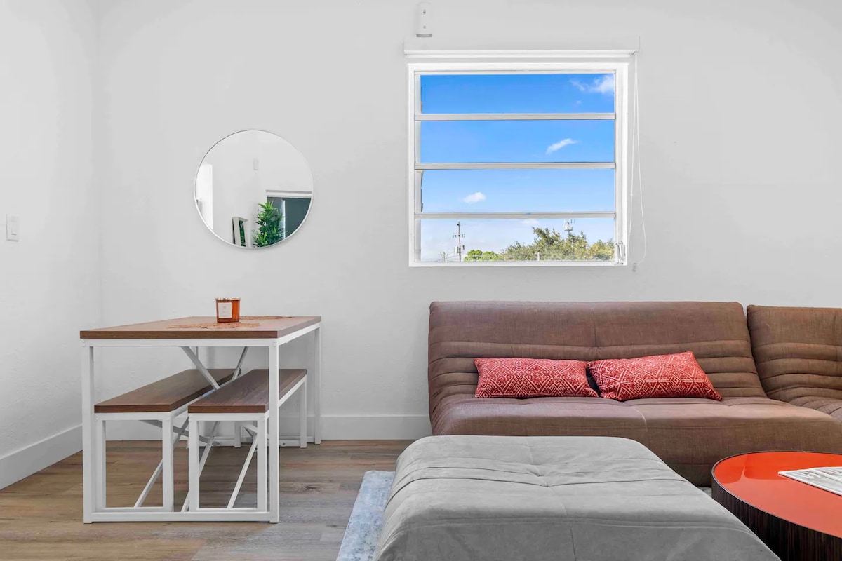 A bright living space features a comfortable sectional couch, positioned near a window allowing natural light to fill the room. A round mirror hangs on the wall opposite a dining table with two benches, complemented by a decorative candle.