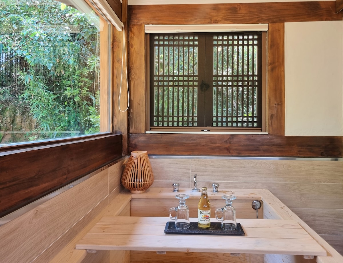 A serene bathing space is featured, highlighting a wooden soaking tub positioned against a window with bamboo lattice. A small wooden table sits beside the tub, adorned with glasses and a beverage, inviting relaxation amidst the natural greenery visible outside.