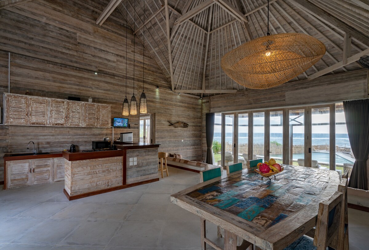 An open-plan living area is highlighted by high cathedral ceilings and wooden walls. A large dining table with an eclectic design occupies the center. The kitchen features modern appliances, and large glass doors provide a view of the ocean and access to the outdoor space.