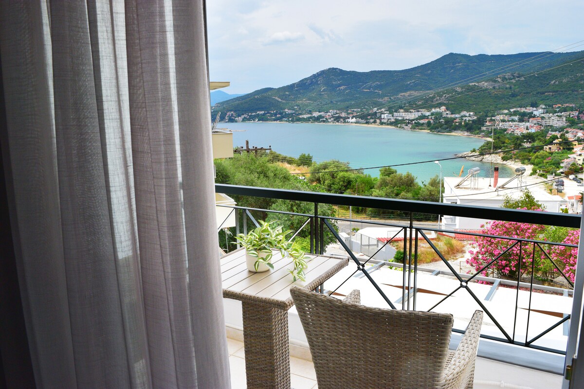 A balcony offers sweeping views of the Aegean Sea and surrounding hills, framed by partially drawn light curtains. A small table and a wicker chair create an inviting space for relaxation while enjoying the scenery.