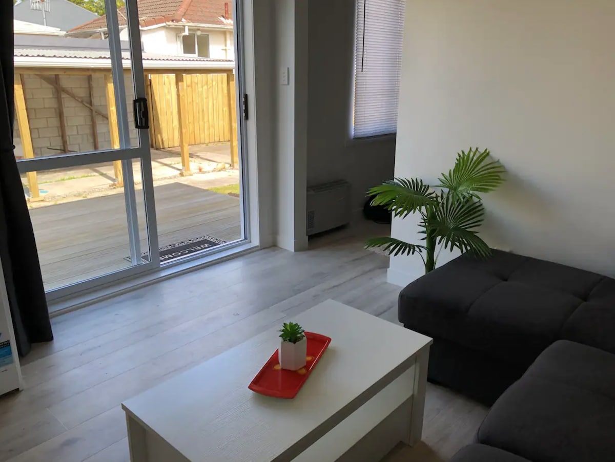 A light-filled living space features a gray sofa paired with a simple coffee table, adorned with a small potted plant and a decorative tray. Floor-to-ceiling glass doors open to a private deck, with a lush green view visible through the partially drawn curtains.