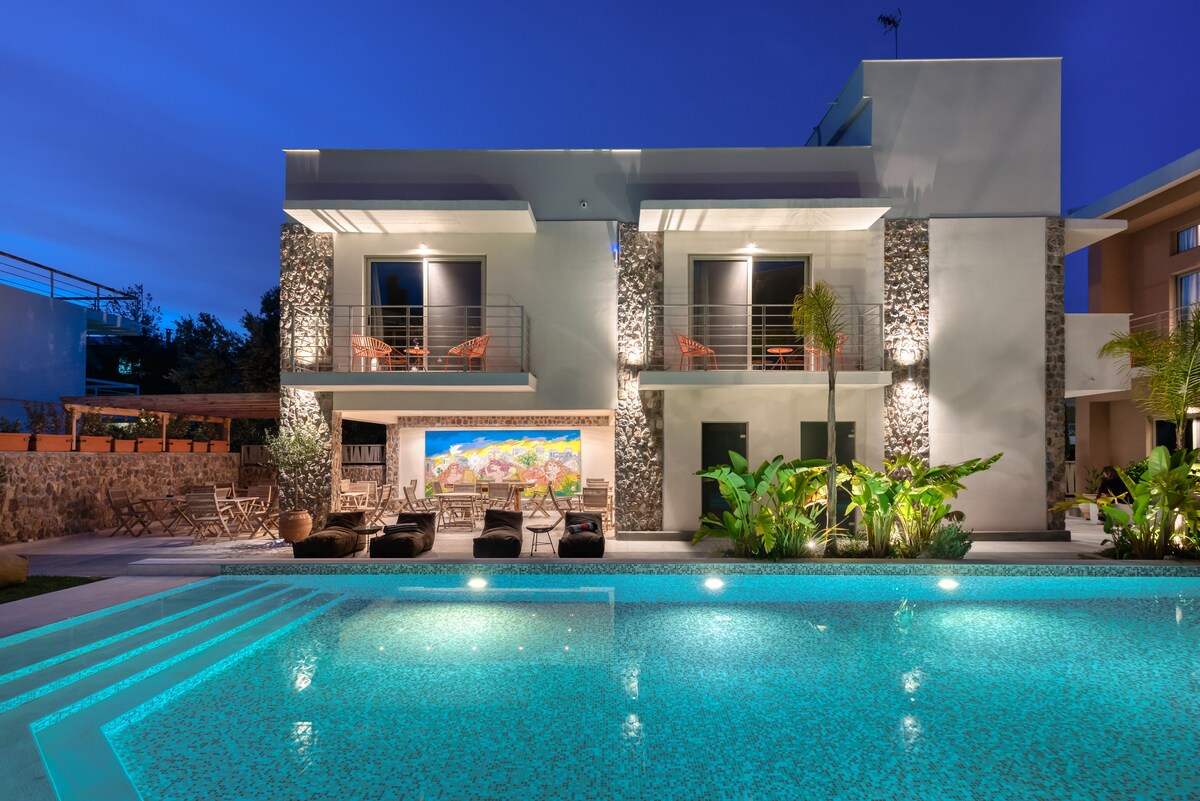 The modern exterior of the property showcases a sleek design with stone accents. A vibrant pool is illuminated in hues of blue, reflecting the surrounding greenery and outdoor seating. The two levels feature large windows and private balconies for views of the lush landscaping.