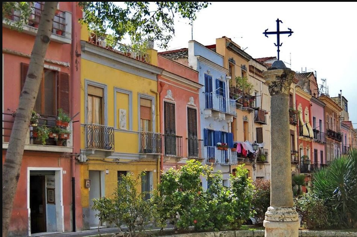 A row of colorful buildings with balconies adorned with plants and hanging laundry can be seen. The central stone column features a cross at the top, surrounded by greenery. The vibrant facades reflect a blend of history and charm in a lively urban setting.