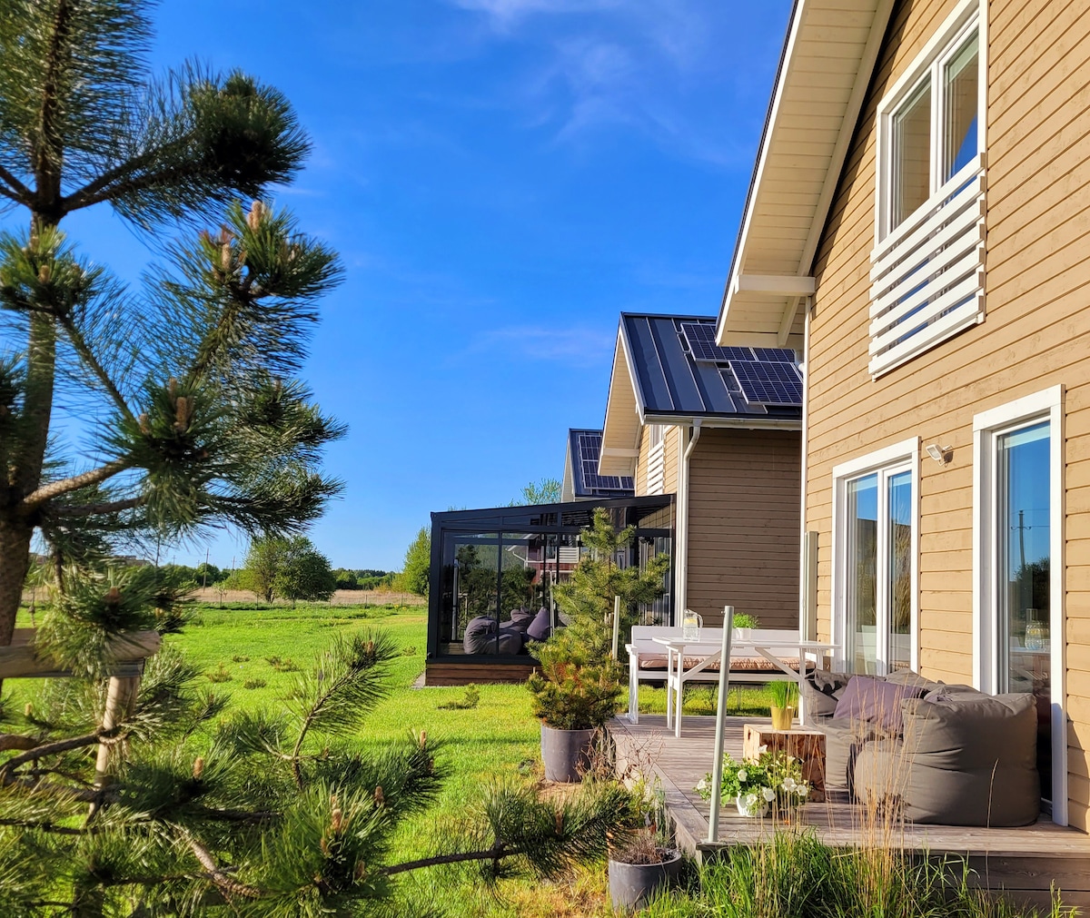 A light brown wooden house features large windows, allowing natural light to fill the interior. A patio area with comfortable seating and plants is visible, set against a backdrop of green fields and a clear blue sky. Solar panels are installed on the roof.