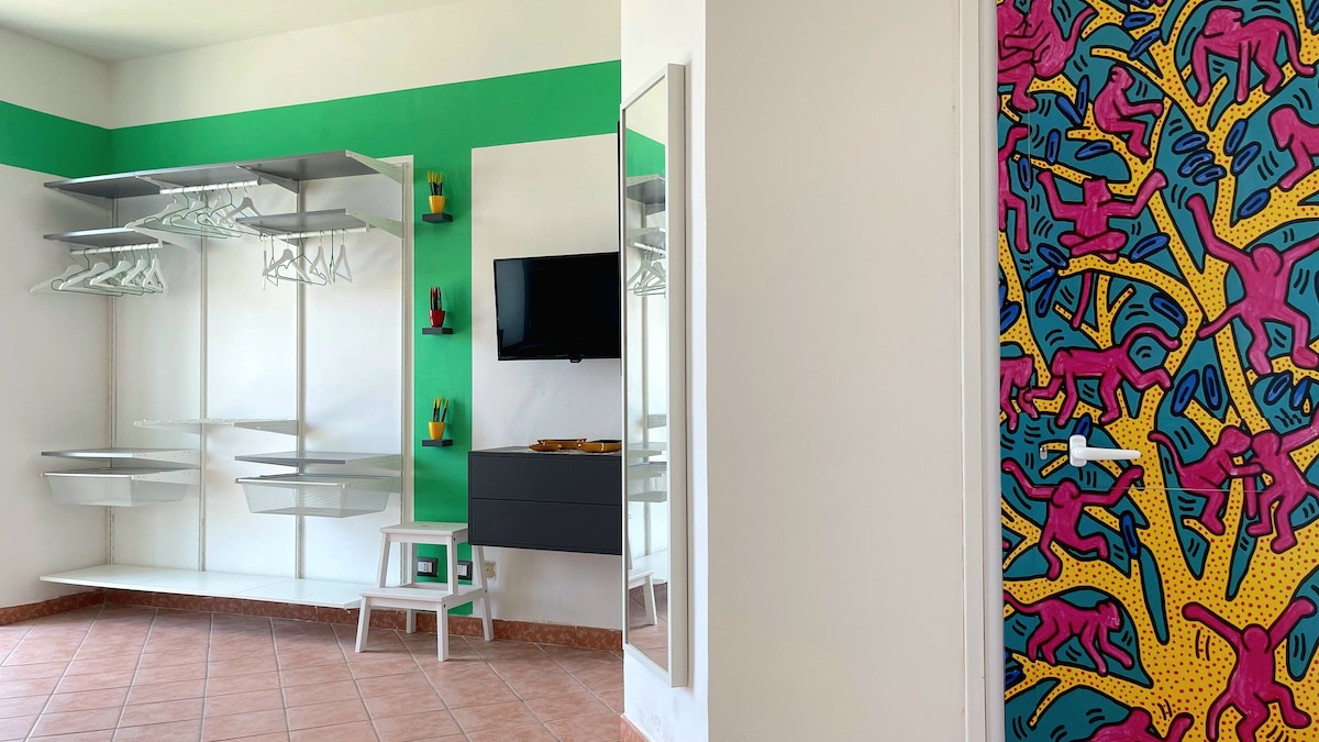 A bright and functional living space is displayed with vivid green accents. Shelving units for storage are seen on one wall, complemented by a mirror and a television. A colorful door adorned with artwork adds a playful touch, enhancing the room's creative atmosphere.