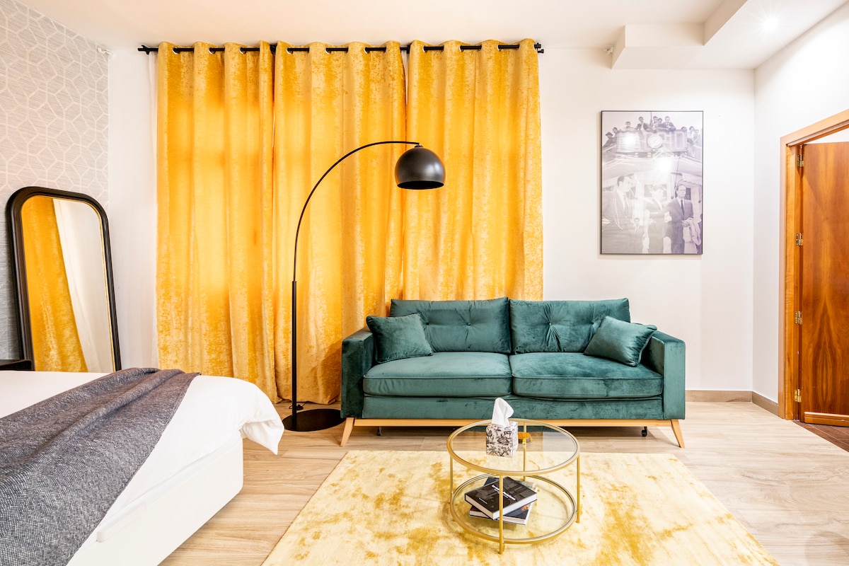 A spacious living area features a vibrant yellow curtain backdrop, providing a lively contrast to the teal sofa. A round glass coffee table sits on a soft area rug, complemented by a stylish floor lamp. The overall layout ensures a comfortable and inviting atmosphere.
