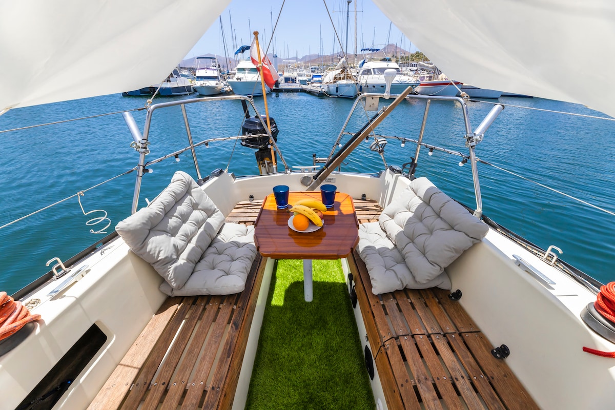 An inviting seating area on the deck is arranged with two cushioned benches and a wooden table. Bright blue mugs and a banana are placed on the table. The view extends to a tranquil marina filled with moored boats and clear blue water.