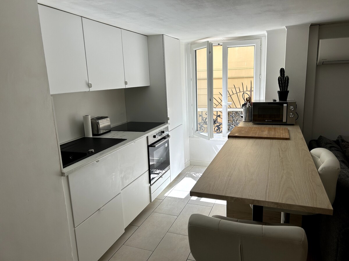 A well-equipped kitchen features modern white cabinetry and a sleek black countertop. A comfortable dining table with light-colored wood provides seating for two. Natural light floods the space through a pair of windows, enhancing a bright and airy atmosphere.
