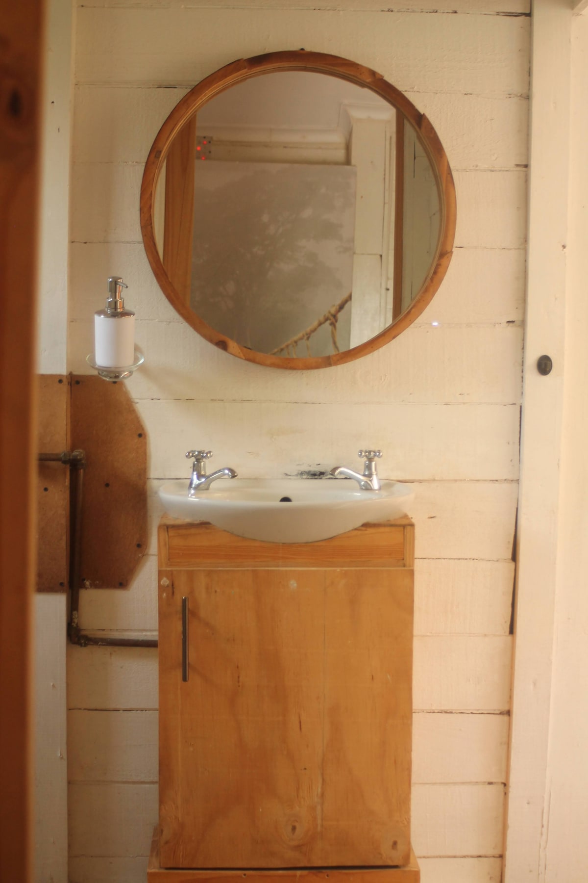 A simple bathroom is featured, showcasing a circular mirror above a wooden sink cabinet. The cabinet is crafted from light-colored wood, and the sink is equipped with two polished metal faucets. Soft light reflects off the mirror, illuminating the rustic wooden walls.