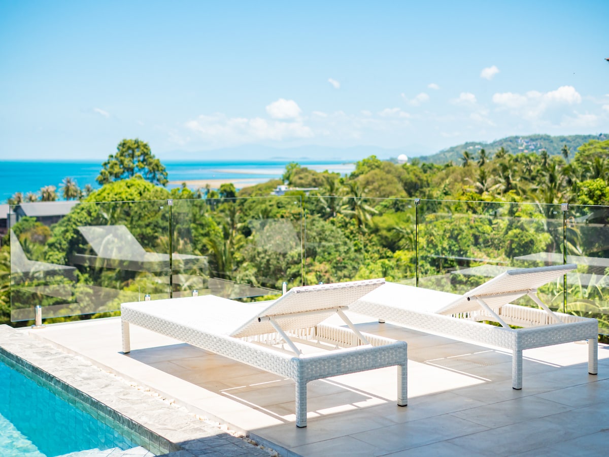 Two sun loungers are positioned by a clear blue pool, offering views of the lush greenery and the distant sea. The clean lines of the loungers reflect a modern design, while the surrounding landscape enhances the serene outdoor setting.
