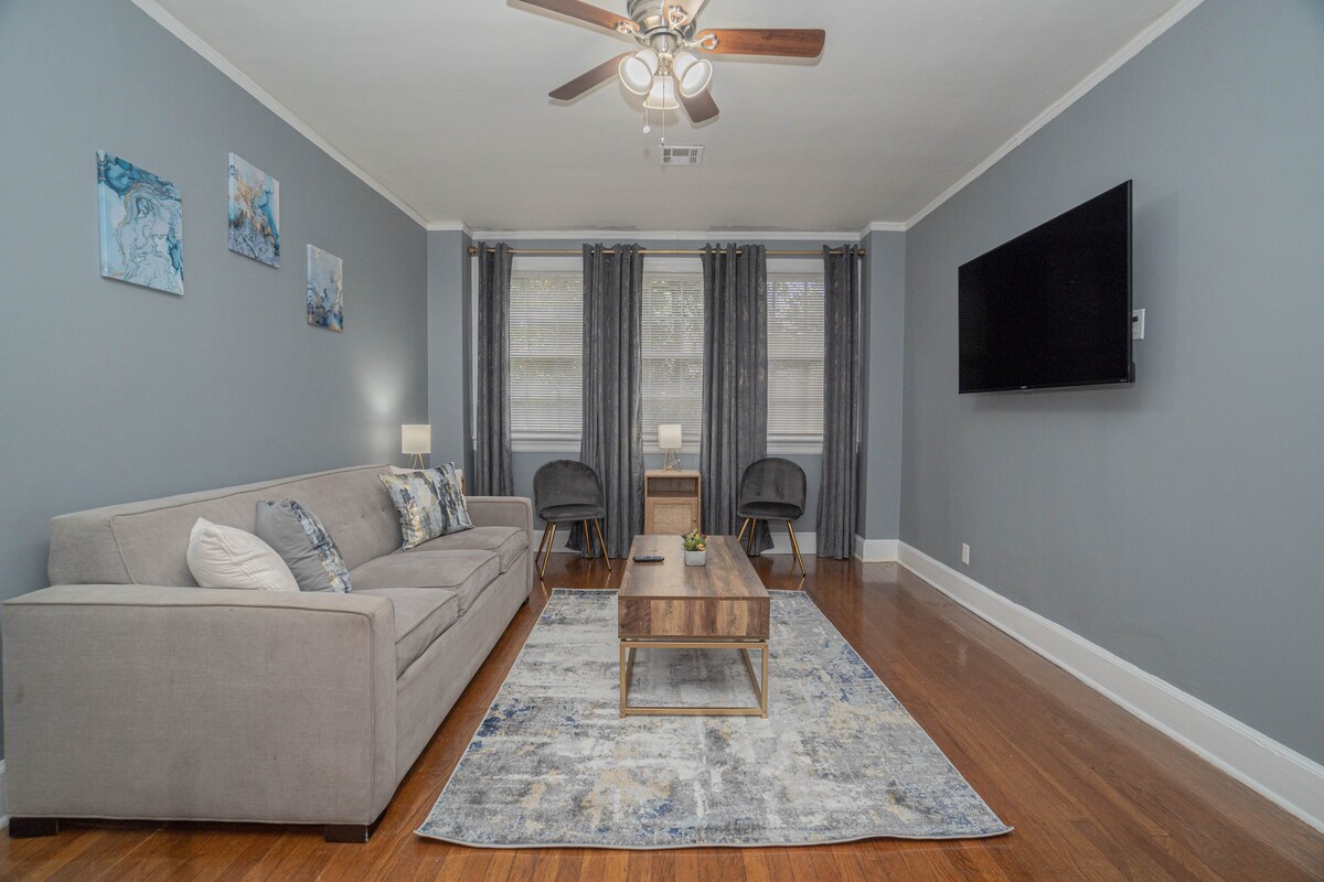 The living area features warm wooden flooring and a spacious sofa, complemented by a coffee table. A Smart TV is mounted on the wall, and two chairs await near a small plant. Soft gray walls and curtains enhance the bright ambiance of the room.