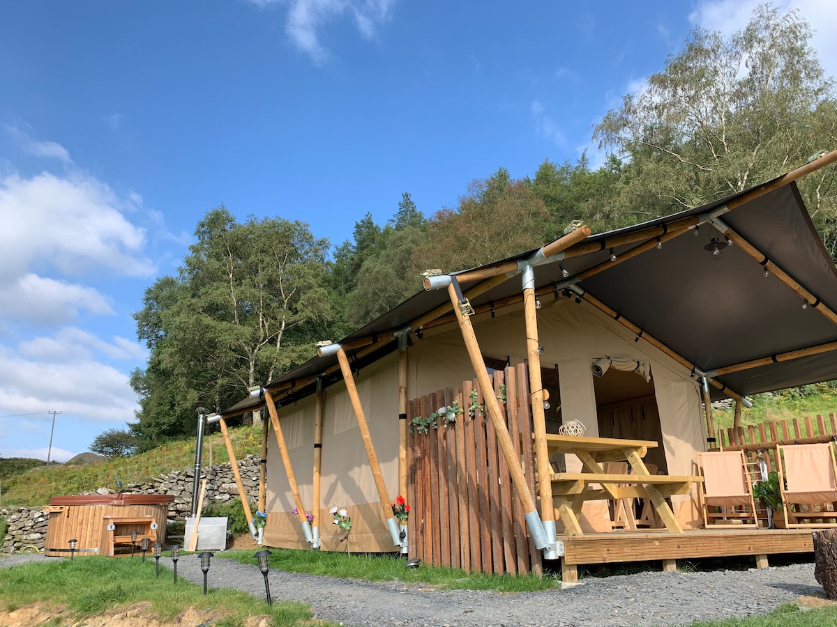 The Safari Lodge is situated amidst lush greenery, featuring a canvas exterior with wooden supports. A large, covered porch includes a picnic table and two wooden chairs. Nearby, a wood-fired hot tub sits invitingly on pebbled ground, surrounded by scenic countryside.