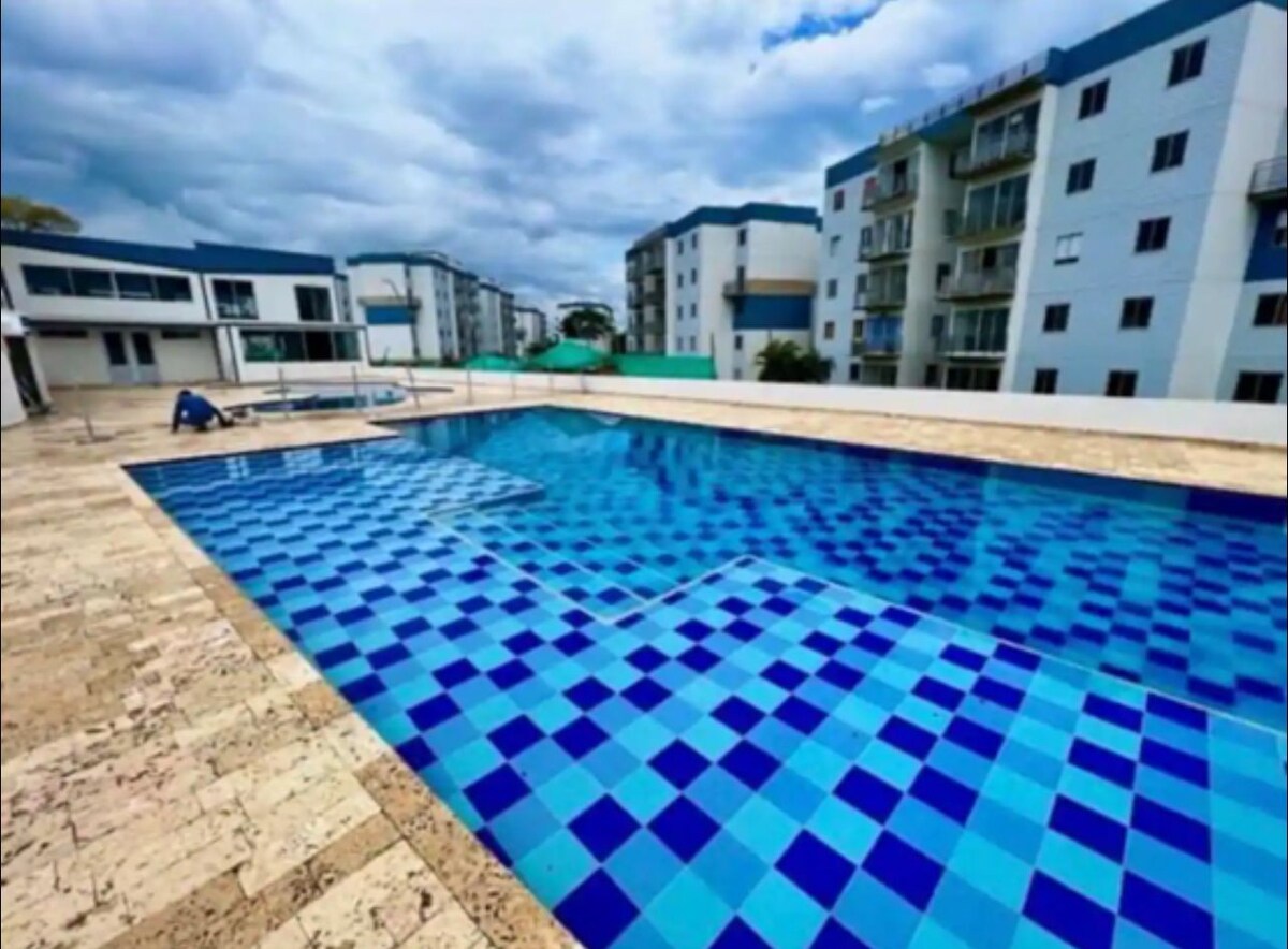 An expansive swimming pool is shown, featuring a vibrant blue and white tiled pattern. Surrounding the pool, a spacious deck offers ample lounging area, with comfortable seating visible nearby. Multiple buildings are seen in the background, creating a serene environment for relaxation.