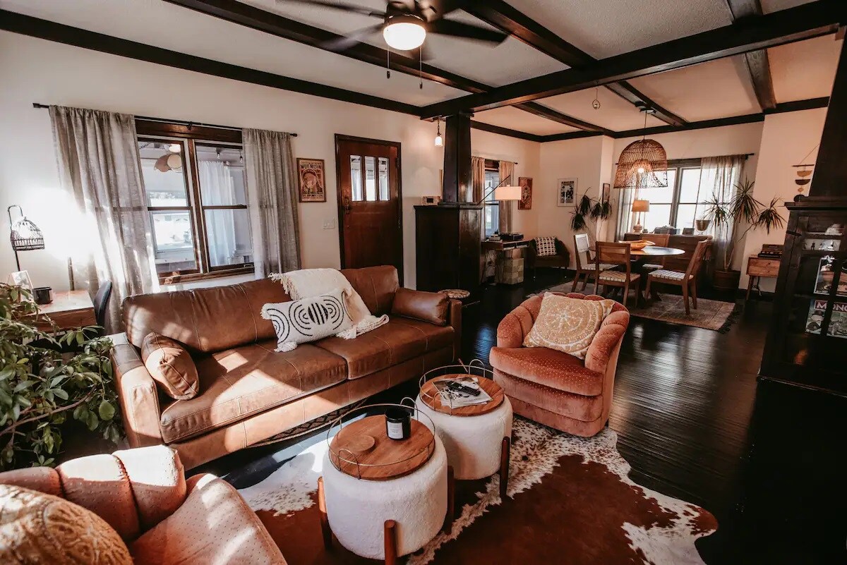 A comfortable living area features a leather sofa and plush armchairs arranged around a circular coffee table. Natural light floods the space through large windows, highlighting the wooden beams and tasteful decor. A dining area with a wooden table is visible in the background.