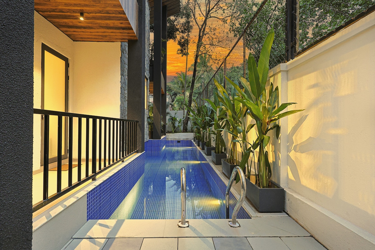 A private swimming pool is framed by lush greenery and potted plants, creating a serene atmosphere. Tiles in shades of blue reflect the warm hues of the sunset sky. The entrance to the villa is visible, along with a wooden awning overhead.