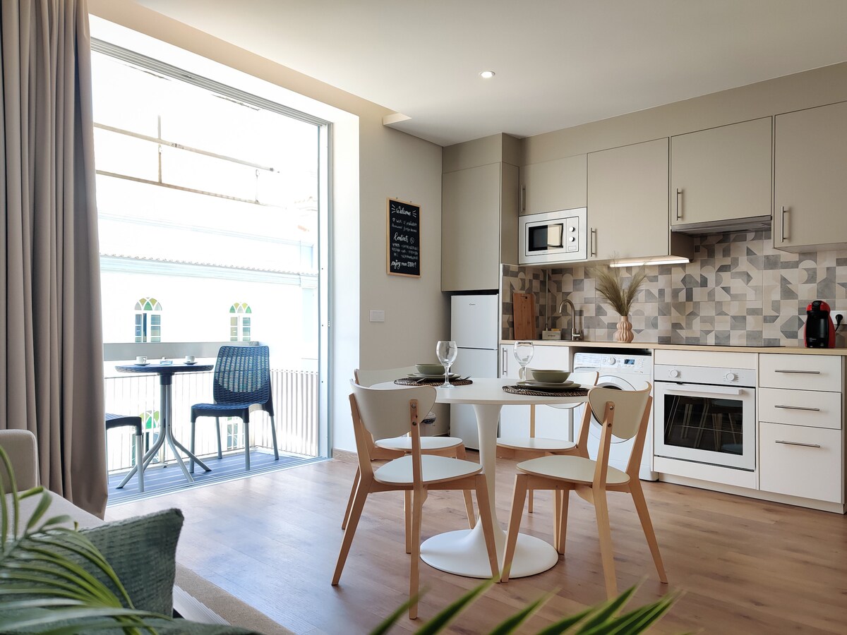 A modern kitchen and dining area are showcased, featuring a round table with four white chairs. The kitchenette is equipped with a stove, microwave, and decorative backsplash. A large window allows natural light, revealing a balcony with an outdoor table and chairs.