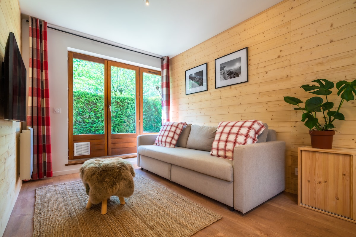 A welcoming living area features a light gray sofa adorned with red-checkered pillows, complemented by a natural area rug. Large glass doors provide views of a lush outdoor space, while wooden paneling and framed black-and-white photographs enhance the cozy atmosphere.