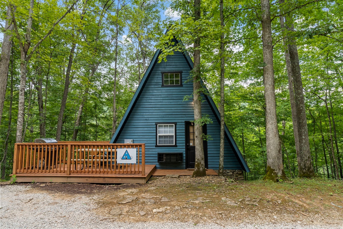 An A-Frame cabin with blue siding is surrounded by tall trees, offering a sense of seclusion. A wooden deck extends from the entrance, providing a welcoming space to relax. The gravel path leads up to the cabin, enhancing its rustic charm.