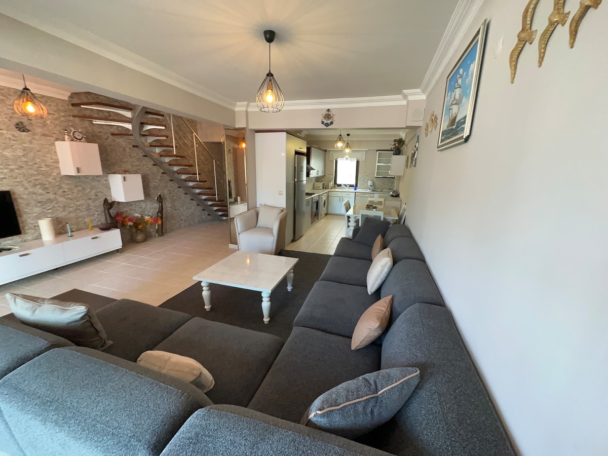 A spacious living area is shown, featuring a large grey sectional sofa with decorative pillows. A white coffee table is positioned in front of the sofa, while a staircase leads to the upper level. Natural light fills the space, creating a welcoming ambiance.