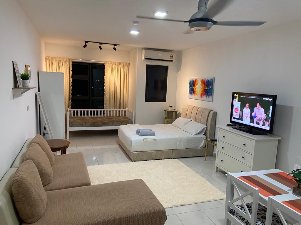 A spacious studio living area is presented, featuring a queen bed and a day bed with soft linens. A beige sofa sits against a light-colored rug, while a television is mounted on a white dresser. Natural light illuminates the space through large windows with sheer curtains.