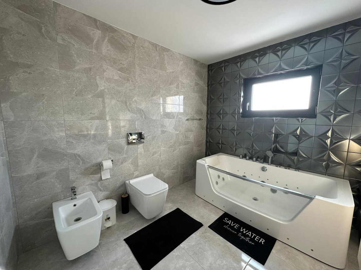 A modern bathroom features a sleek, freestanding jacuzzi tub and a bidet. The walls are adorned with stylish gray tiles, and a large window provides natural light. A black bath mat with text adds contrast to the light tile flooring.