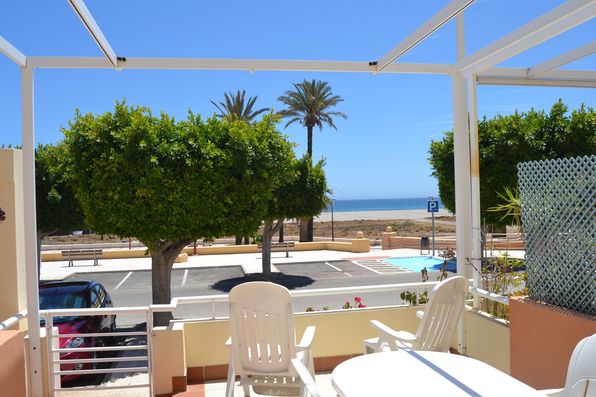 A shaded outdoor porch area features a table and chairs, providing a view of palm trees and the beach. The sea is visible in the distance, complemented by a clear blue sky. The surrounding area includes parking spaces and landscaped greenery.