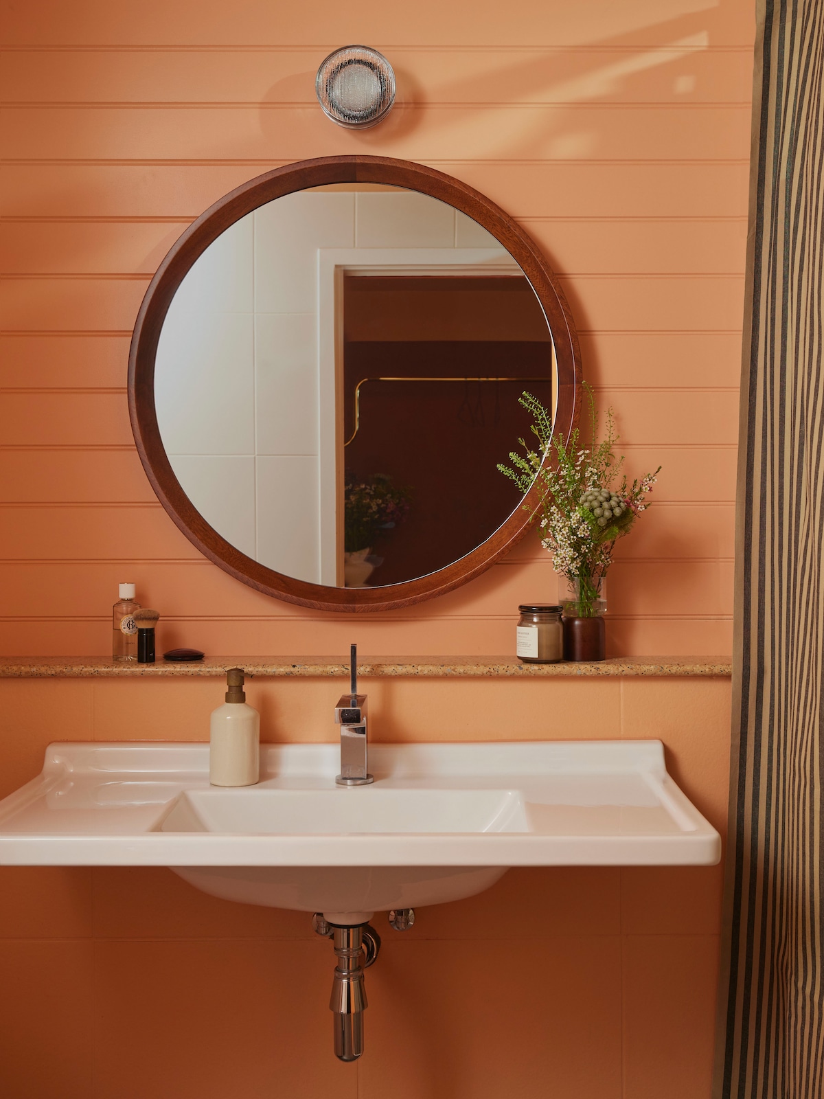 A contemporary bathroom features a round mirror above a sleek sink. The warm peach-colored walls are complemented by a countertop adorned with minimalist toiletries and a small vase of greenery. A striped shower curtain adds a touch of texture to the space.