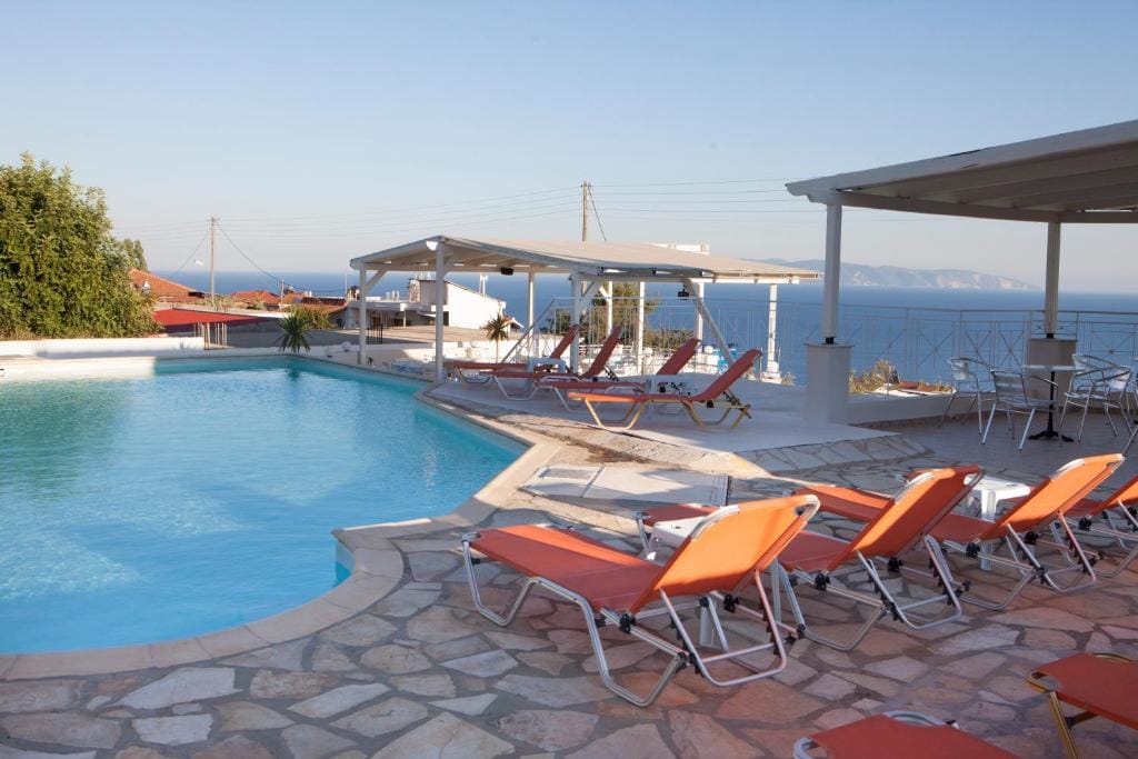 A spacious pool area is viewed, featuring a turquoise pool surrounded by sun loungers. The loungers are positioned to face the sea, with a pergola providing shade nearby. Clear blue skies and distant hills are visible in the background.