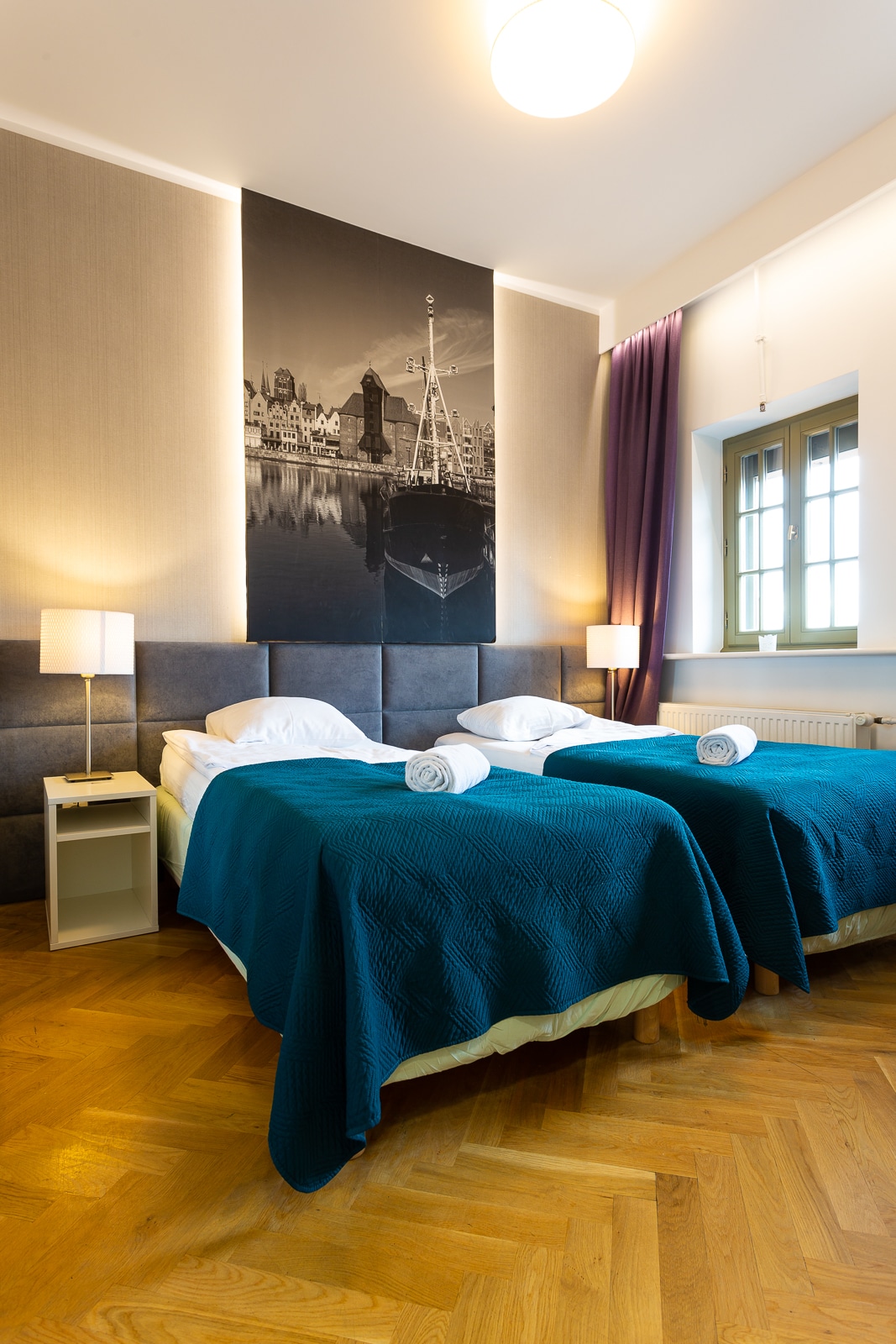 The image displays a guest room featuring twin beds, each adorned with a light blue blanket and white towels. A large black-and-white photograph of a cityscape enhances the wall behind the beds. Soft lighting from bedside lamps creates a comfortable ambiance.