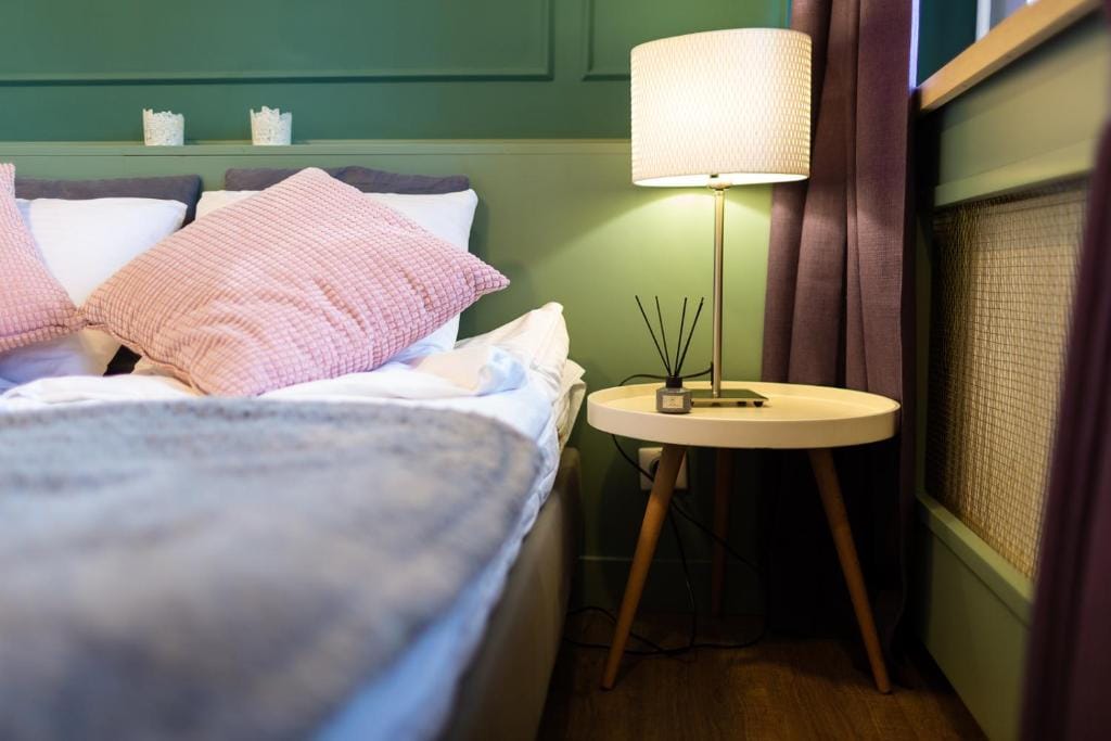 A cozy sleeping area is presented with a spacious bed adorned with soft pillows and a warm blanket. A bedside table hosts a stylish lamp and an aromatic diffuser, with muted green walls creating a calm environment. Natural light filters in through a nearby curtain.
