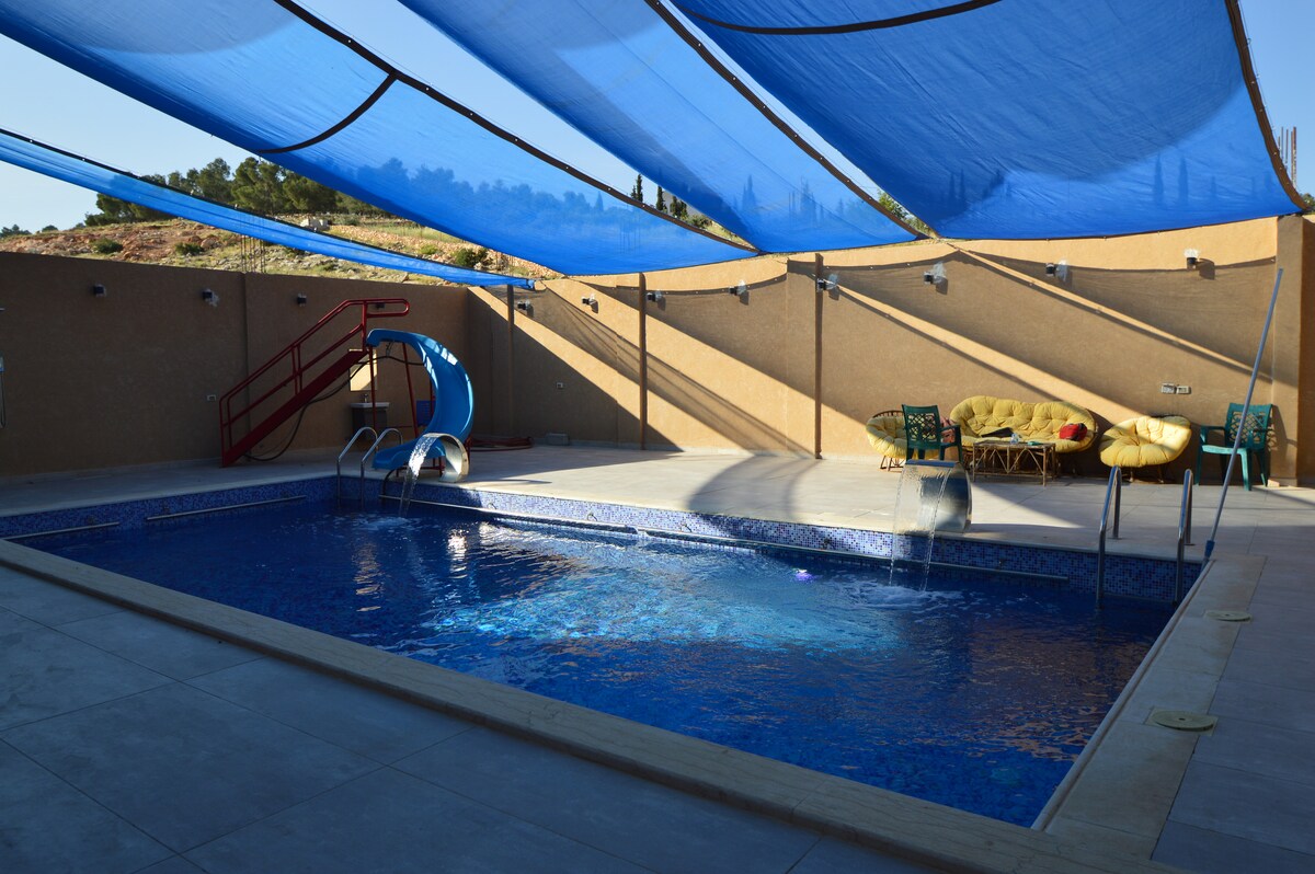 A swimming pool measuring 10 meters long and 5 meters wide is visible, featuring a slide and two waterfalls. The pool area is shaded by a blue canopy. Chairs and loungers are positioned nearby for relaxation.