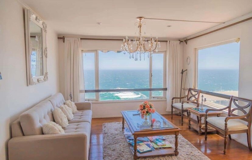 A light-filled living area offers sweeping views of the ocean through large windows. A comfortable sofa is positioned across from a wooden coffee table, adorned with a floral arrangement. Natural light enhances the space, complemented by elegant drapes and a stylish chandelier.