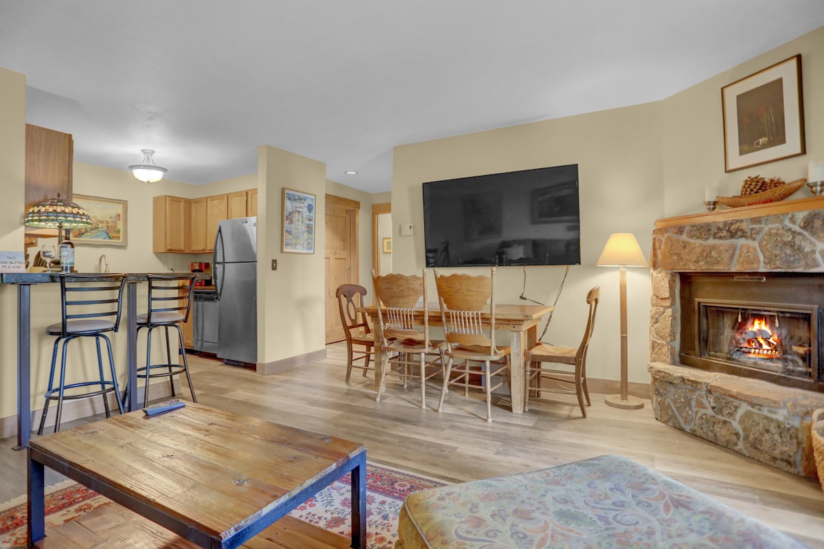 The spacious living area features a dining table with six wooden chairs and a cozy seating arrangement. A flat-screen TV is mounted on the wall, while a warm wood-burning fireplace adds charm. The kitchen area showcases a breakfast bar with two stools, creating a welcoming atmosphere.