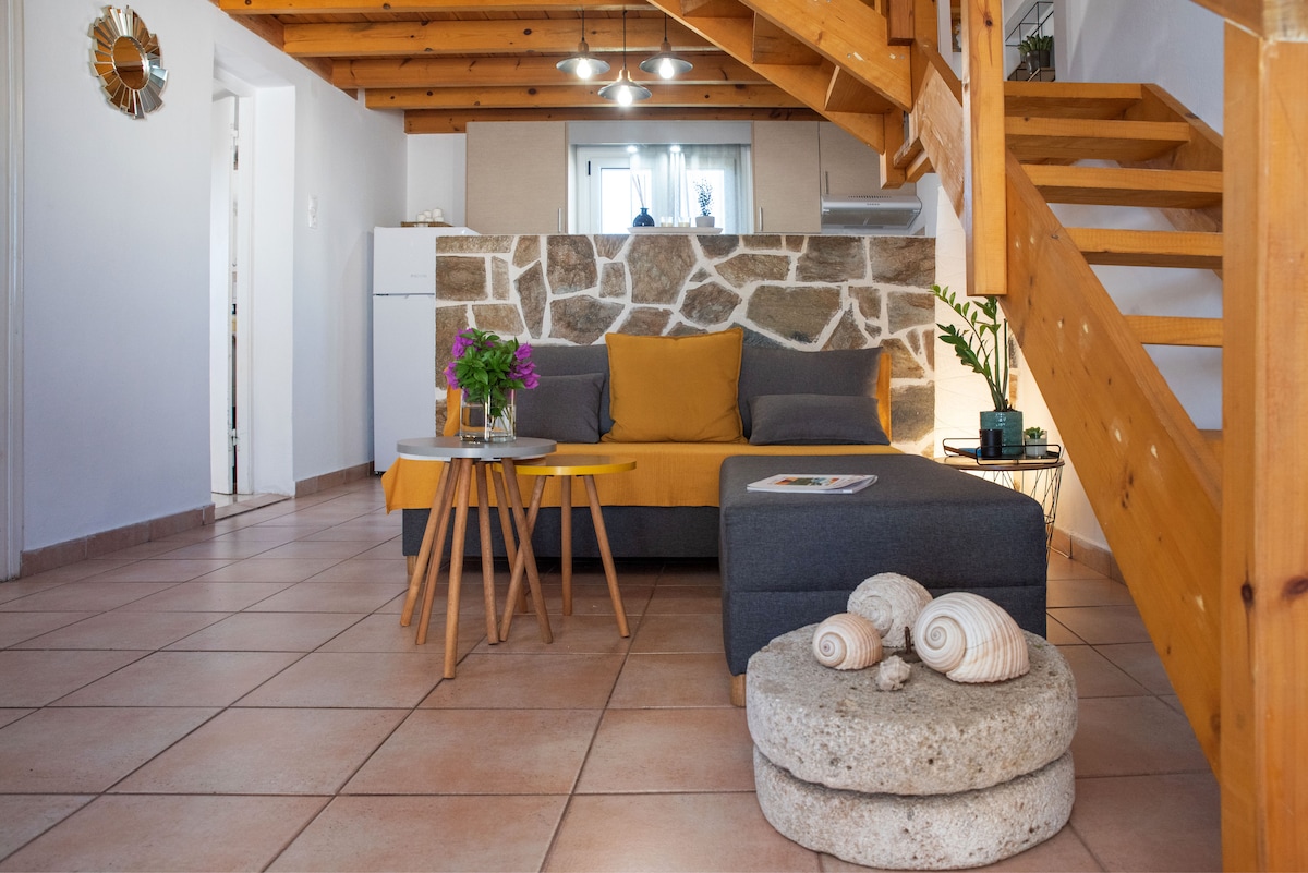 A cozy living area features a gray sofa and a wooden coffee table, accompanied by a round stone ottoman. Natural light fills the space, highlighting the stone wall and wooden beams above. A small vase of flowers adds a touch of color.