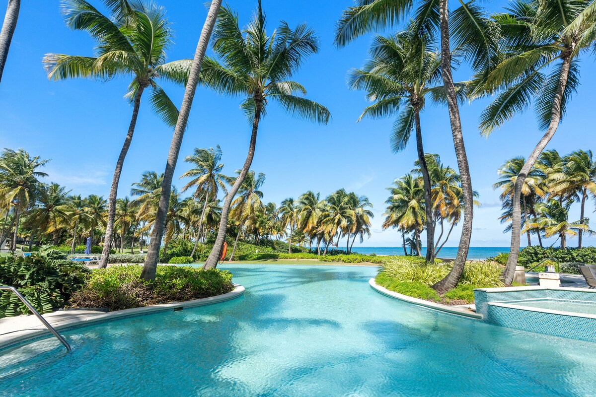 A serene infinity pool is surrounded by lush palm trees, leading the eye toward the sky and ocean beyond. The clear blue water reflects the tropical landscape, creating a peaceful ambiance ideal for relaxation.