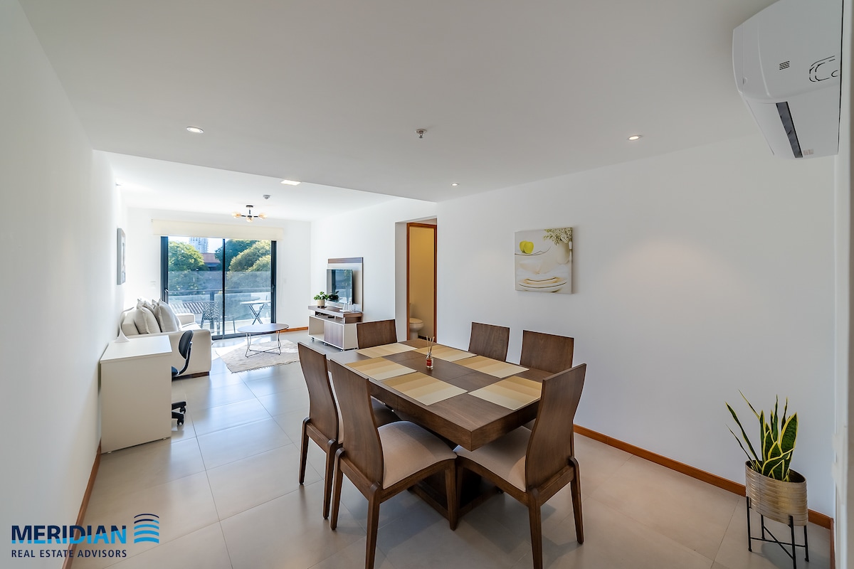 A modern dining area features a rectangular table surrounded by six wooden chairs. A large window provides natural light, revealing a view of the outdoors. Wall art adds a subtle design element, while neutral tones and minimalist decor create a spacious and uncluttered environment.