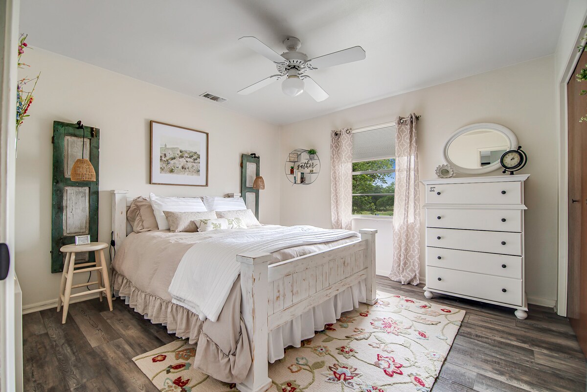 The airy bedroom features a spacious bed adorned with neutral linens, complemented by decorative pillows. A dresser and a side table provide functional storage. Natural light filters through sheer curtains, and a floral area rug adds softness to the wooden floor.
