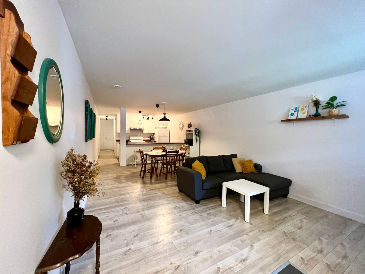A spacious living area features a comfortable gray sofa with accent pillows, a small white coffee table, and a decorative wooden end table. Light wood flooring complements the neutral walls. A dining area with a wooden table and chairs is visible in the background, alongside a fully equipped kitchen.