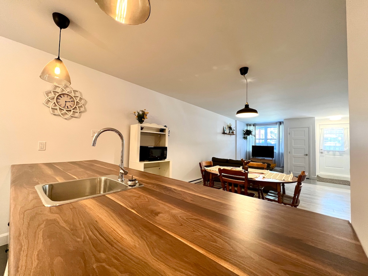 A wooden counter with a sink is positioned in the foreground. Soft light is provided by hanging pendant lights above. In the background, a dining area with a table and chairs is visible, along with a cozy living space featuring a television and well-placed decor.