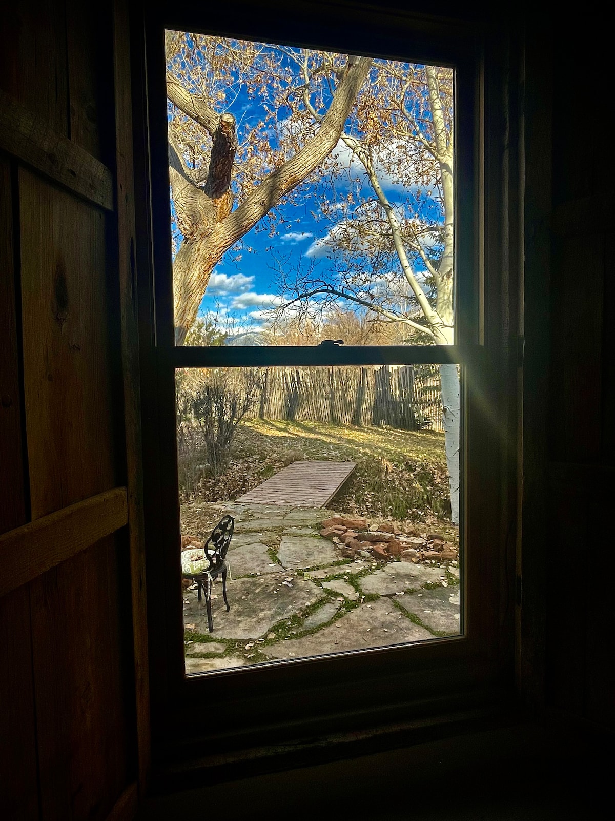 A window frame offers a view of a path made of stone leading through a serene outdoor space. A small table with a chair is visible, surrounded by trees and open sky, conveying a peaceful connection with nature.