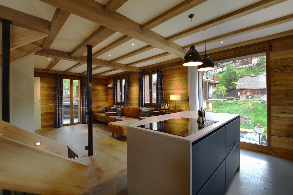 An open-plan living space is highlighted by exposed wooden beams and large windows offering views of the outdoors. A modern kitchen island contrasts with the warm wood walls, while leather sofas provide a comfortable seating area. Natural light filters through, enhancing the inviting ambiance.