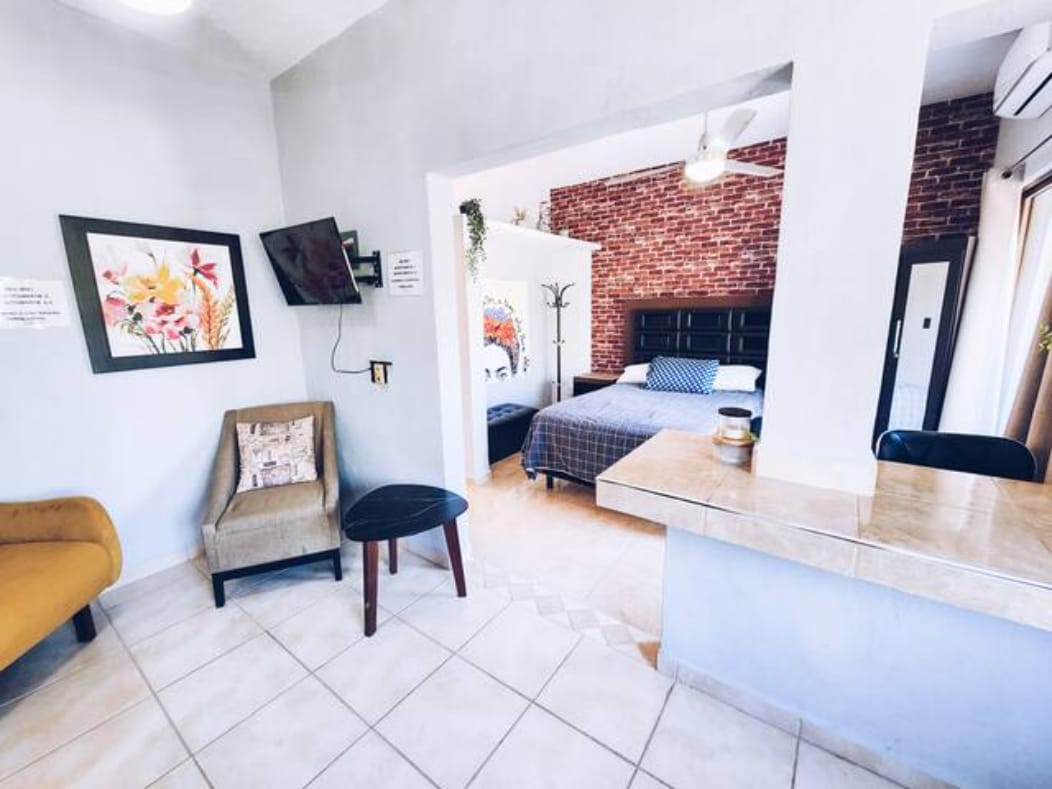 A cozy studio space is presented, featuring a mix of modern and rustic elements. A brick wall is highlighted, with comfortable furniture consisting of a chair and a small table visible in the foreground. A kitchenette area is located to the right, while a spacious bed is seen in the background.