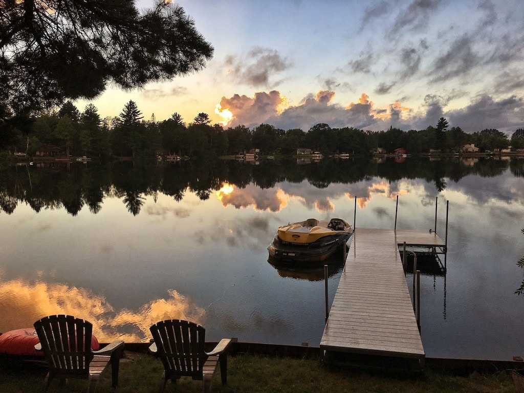 A calm lake is reflected in the early evening light, showcasing gentle hues in the sky. A wooden dock extends into the water, beside a yellow boat. Adirondack chairs are positioned on the shore, offering a relaxed seating area with views of the serene landscape.