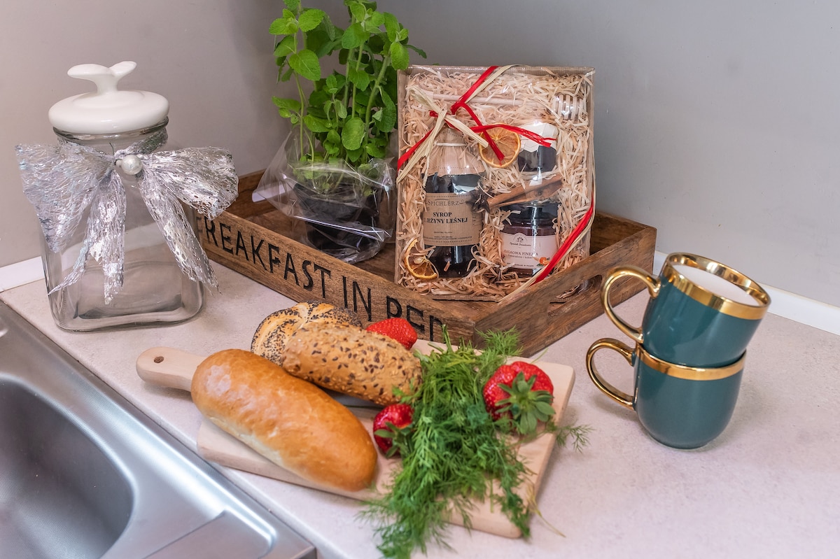 A kitchen counter features a wooden tray holding jars of preserves and a plant. Nearby, a cutting board displays a fresh baguette, strawberries, and herbs. A ceramic mug with a gold band rests beside the food items, creating a welcoming breakfast setup.