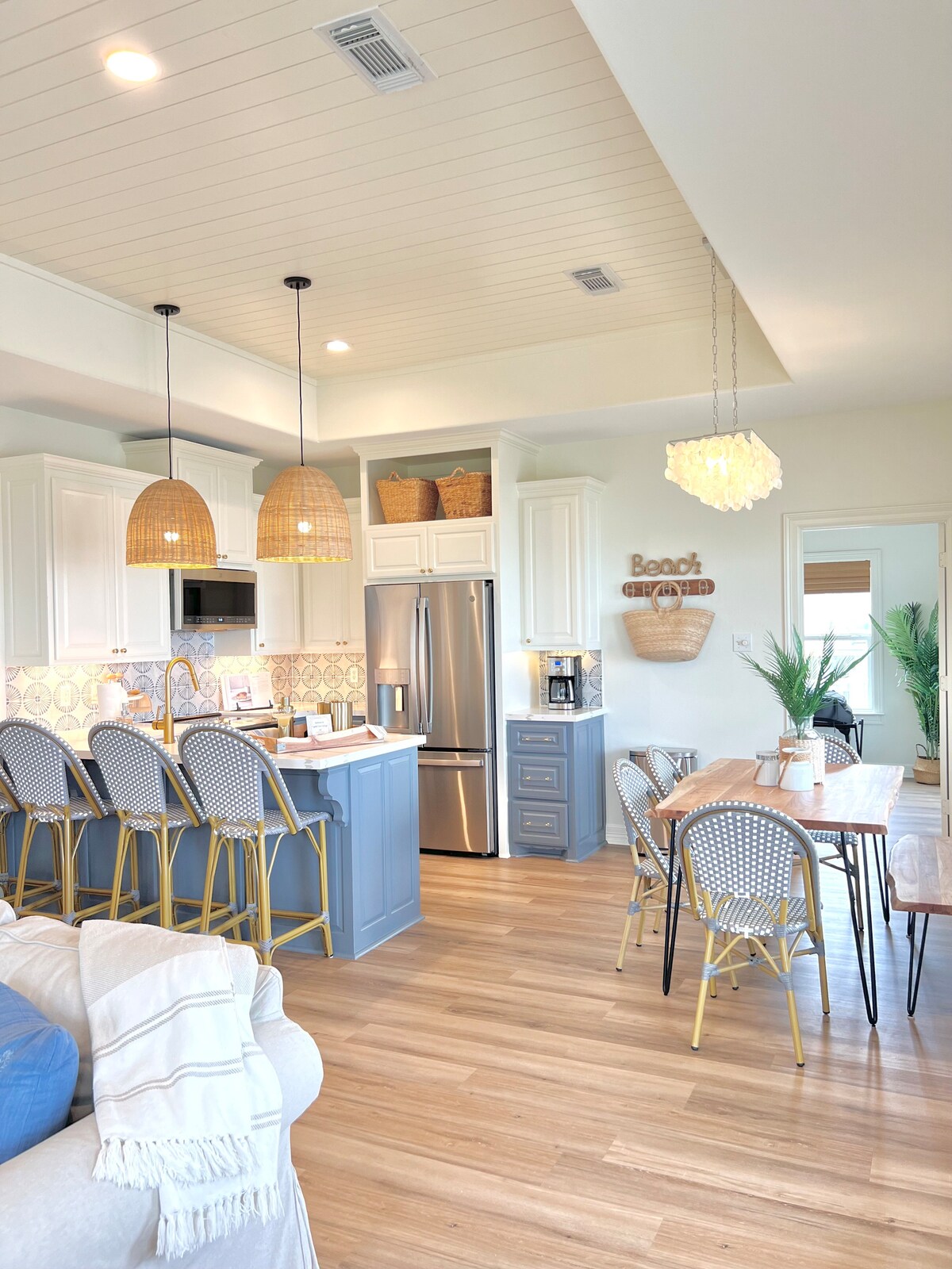 The kitchen features light cabinets paired with a gray island, accented by two woven pendant lights. Four bar stools line the island, while a dining table is set nearby. Sunlight streams through the windows, illuminating the space and highlighting decorative accents.