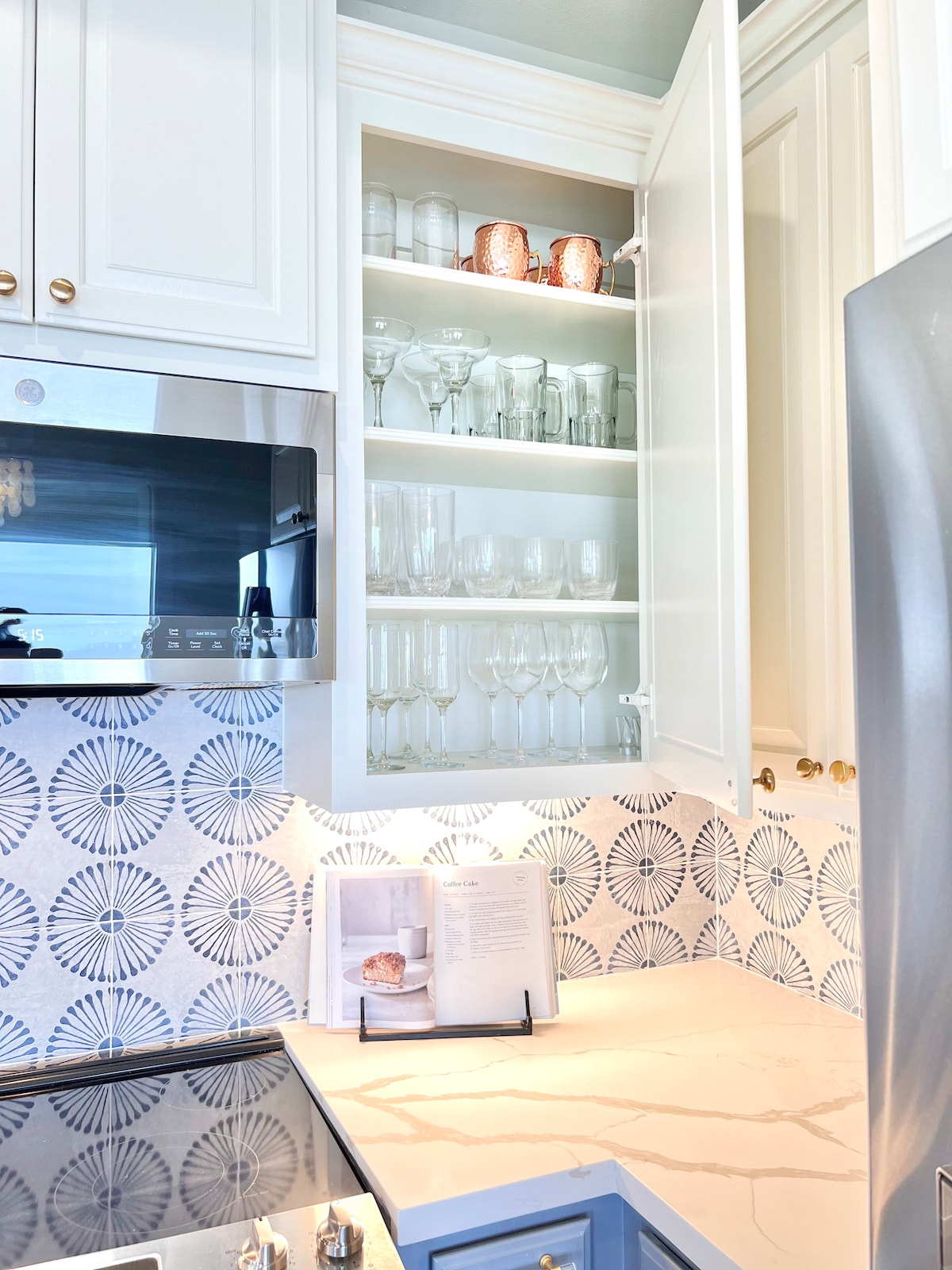 A well-organized kitchen cabinet displays a selection of glassware, including wine glasses and tumblers. Soft lighting highlights the delicate reflections on the glasses. The cabinet features white cabinetry and complements the intricate tile design below, creating a clean and elegant kitchen space.