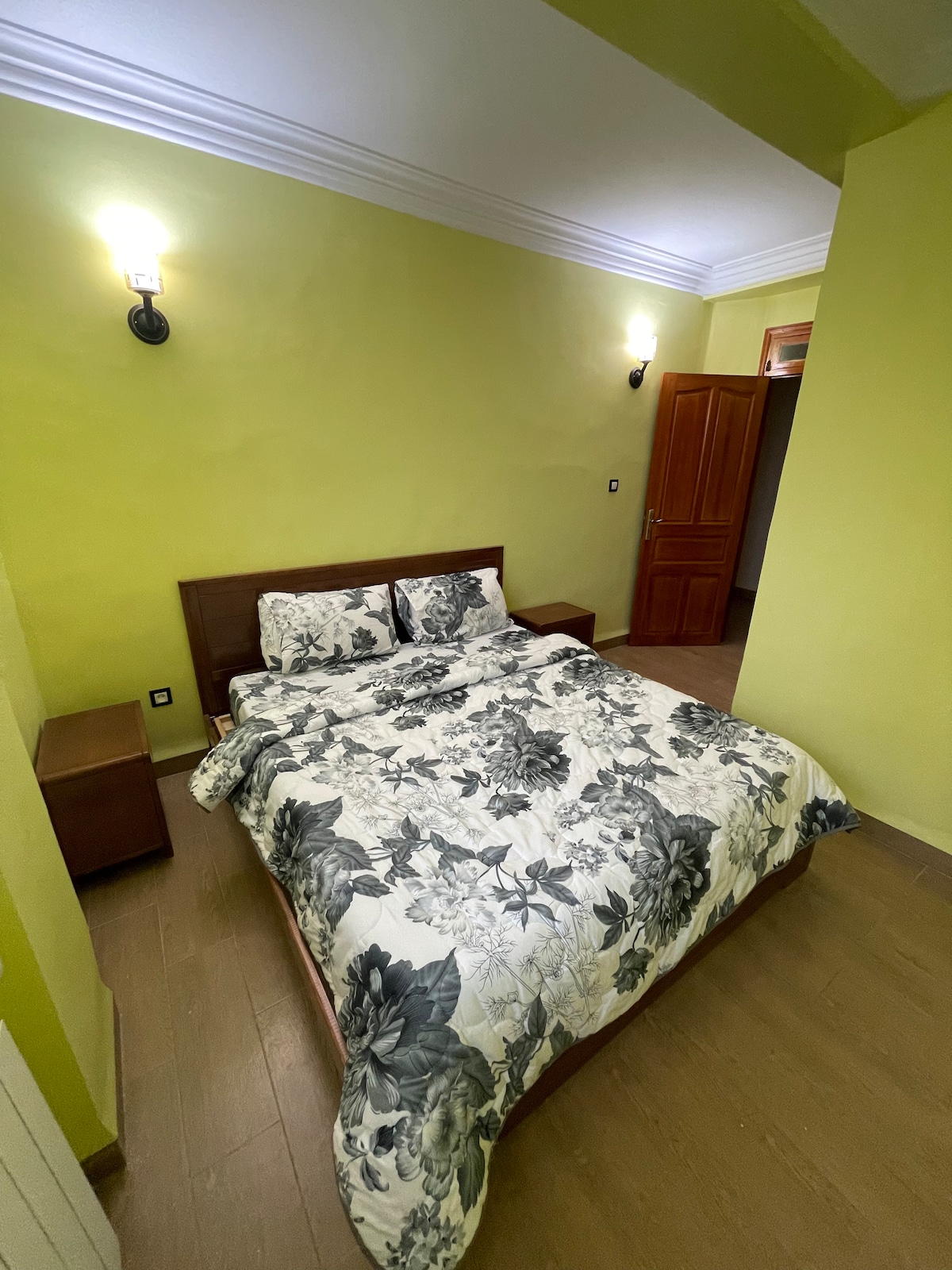 A cozy bedroom features a spacious bed adorned with a floral bedspread, complemented by matching bedside tables. Soft lighting from wall sconces illuminates the green walls, creating a comfortable and inviting ambiance.