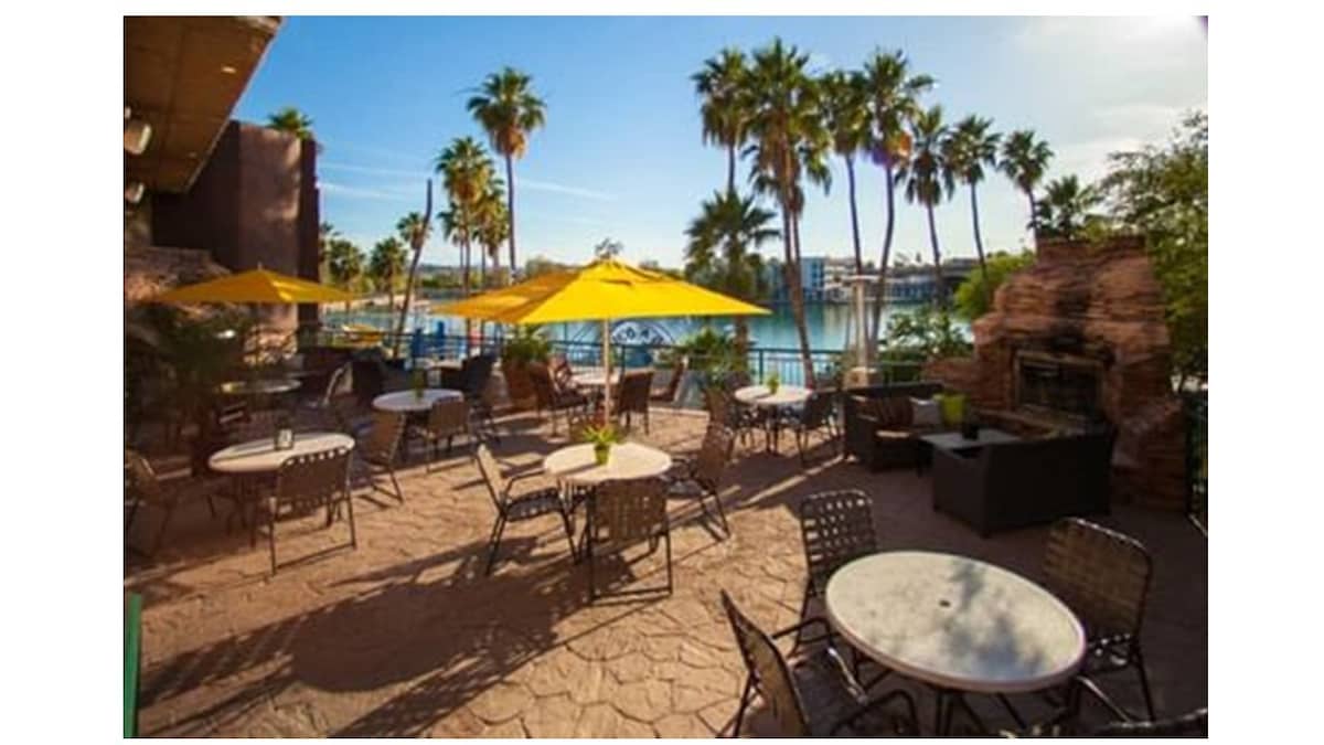 A spacious outdoor seating area features round tables surrounded by metal chairs, shaded by yellow umbrellas. Palm trees are visible in the background, along with a serene waterfront view. A stone fireplace adds a cozy element to the setting.